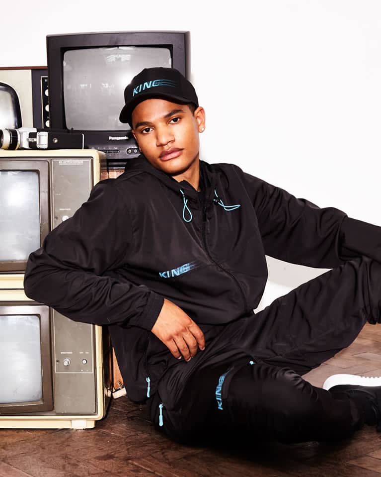 Model wears King Apparel Tennyson curved peak cap and tracksuit in black