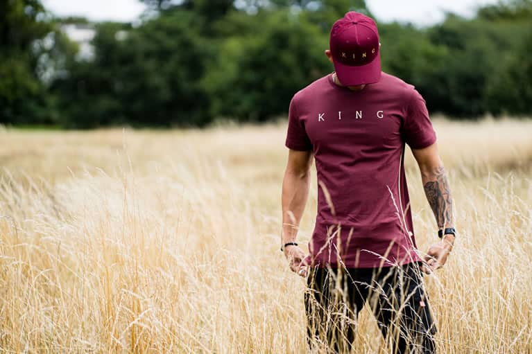 Model wears King Apparel Tennyson Gold curved peak cap and t-shirt in oxblood red