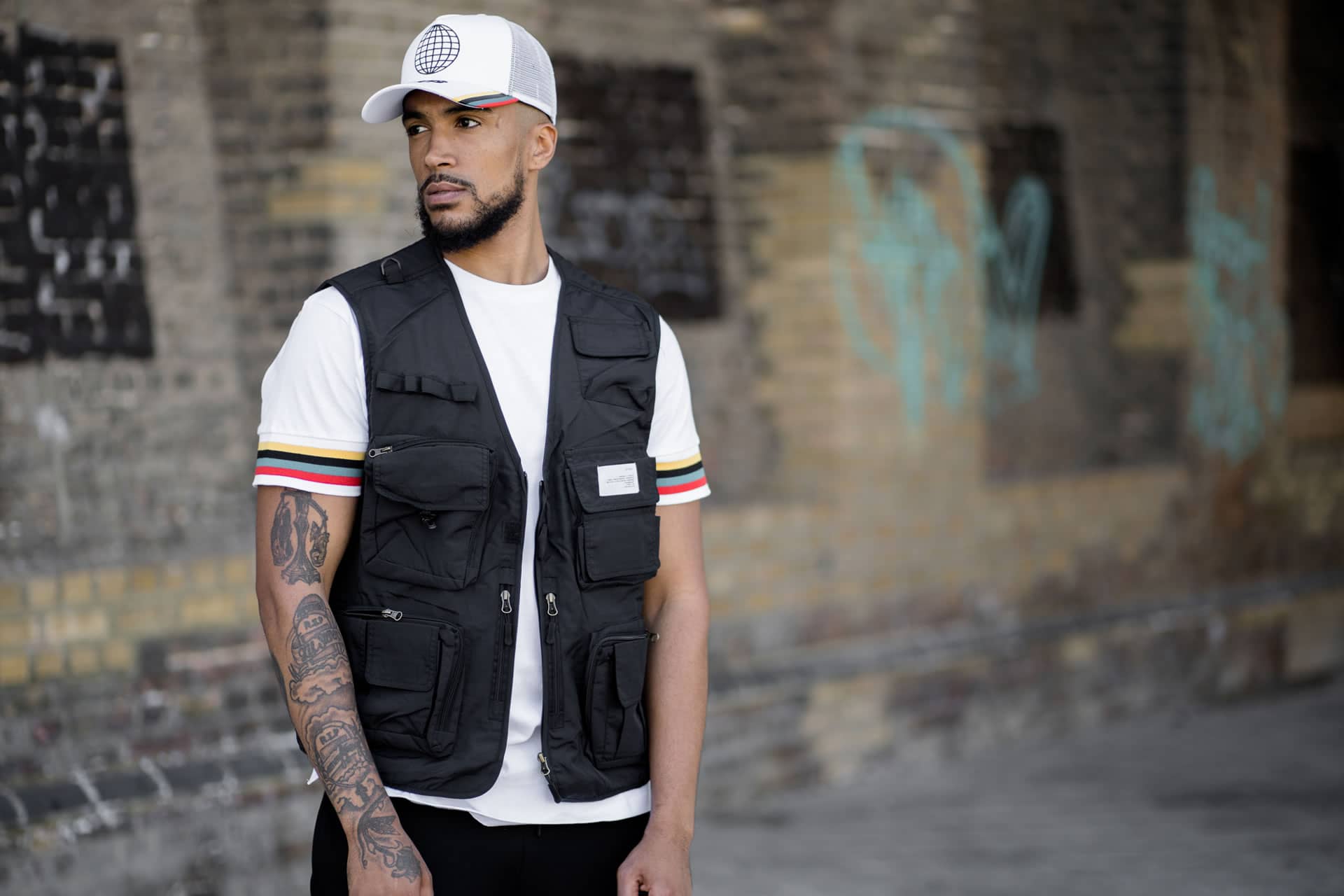Myers wears King Apparel Bethnal mesh trucker cap and t-shirt in white and Earlham Techwear tactical vest in black