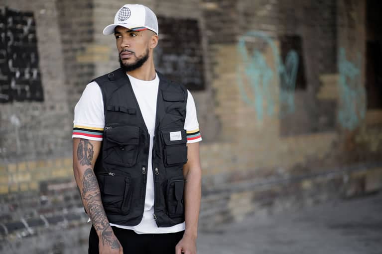 Myers wears King Apparel Bethnal mesh trucker cap and t-shirt in white and Earlham Techwear tactical vest in black